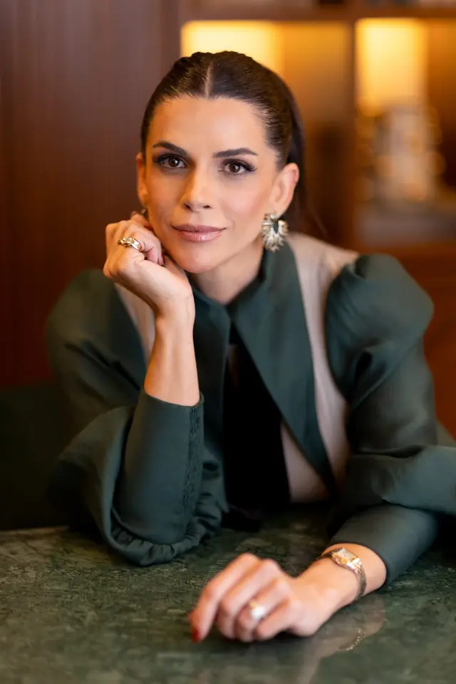 Mel Galhardo no ateliê, de blusa verde, apoiada numa mesa de mármore verde, olhando para a câmera